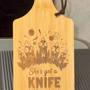 Op de afbeelding: Een bamboe snijplank met een handvat en een leren koord. De plank heeft een gegraveerd ontwerp van cartoon kippen en de tekst "She's got a KNIFE". De plank wordt op een transparante standaard gepresenteerd.