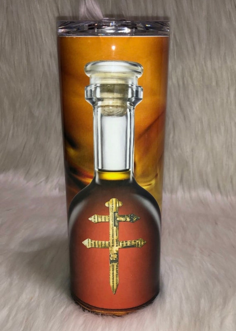 May include: A stainless steel tumbler with a design of a bottle of brown liquor with a gold cross on the label. The bottle has a cork stopper.