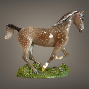 Könnte beinhalten: Eine Keramikpferdefigur in Laufpose. Das Pferd hat eine gesprenkelte braun-weiße Farbe mit einer dunkelbraunen Mähne und einem Schweif. Es steht auf einem grünen Sockel mit Gras und Steinen.