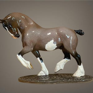 Könnte beinhalten: Keramikpferdefigur in Gehposition. Das Pferd hat einen braunen Körper mit weißen Flecken und schwarzen Beinen mit weißen Hufen. Mähne und Schweif sind schwarz. Die Figur steht auf einem strukturierten Sockel.