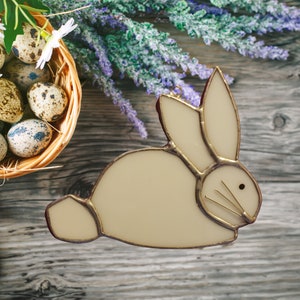 May include: A cream-colored stained glass bunny ornament with a gold-colored outline and whiskers. The bunny is set against a rustic wooden background with a basket of speckled eggs and purple flowers. The ornament is designed for Easter.
