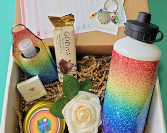 May include: A gift box containing a rainbow glitter water bottle, a Godiva chocolate bar, a rainbow keychain with a heart charm, a white rose, a small candle, and several circular stickers with phrases like Love is Love and Pride.