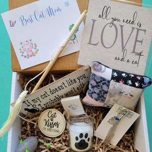May include: A brown cardboard box filled with cat-themed gifts. The box contains a wooden sign that reads 'If my cat doesn't like you, I probably won't either', a white candle with a black paw print, a small gray mouse toy, a white feather keychain with a cat charm, a small white and gray patchwork pillow with a cat design, a white wooden sign that reads 'Cat Kisses Fix Everything', and a white card with the words 'Best Cat Mom' in blue cursive writing.