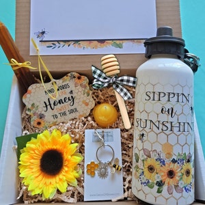 May include: A gift box filled with a variety of items themed around bees and sunflowers. The box includes a honey dipper, a candle, a keychain, a water bottle with the text "Sippin' on Sunshine", and a wooden sign with the text "Kind words are like honey, sweet to the soul".