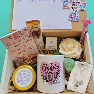 May include: A gift box containing a white mug with the text Choose Joy, a cross-shaped ornament with the text Abide in His Love, a card with the text God has made everything Beautiful, a pen with a pink gem, a candle, a keychain, and bookmarks.