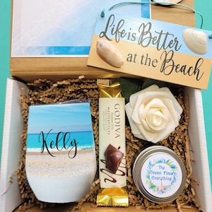 May include: A beach-themed gift box with a brown cardboard box, a white wine tumbler with a beach scene and the name "Kelly" on it, a chocolate bar, a white rose, and a small tin with a beach-themed sticker that says "The Ocean Fixes Everything".