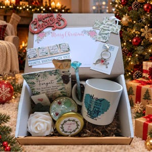 May include: A gift box containing a white mug with blue text, a blue crystal-topped pen, a white artificial rose, a gold-lidded candle labeled Comfort & Joy, a cross ornament, a keychain, magnetic bookmarks, and a card with Merry Christmas text.