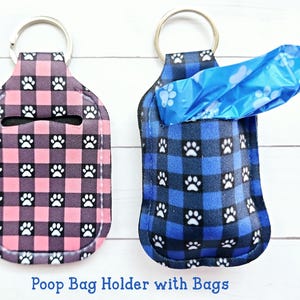 May include: Two dog poop bag holders with paw print and plaid patterns. The holder on the left is pink and black, the holder on the right is blue and black. Both holders have a key ring and a blue poop bag is partially pulled out of the blue holder.