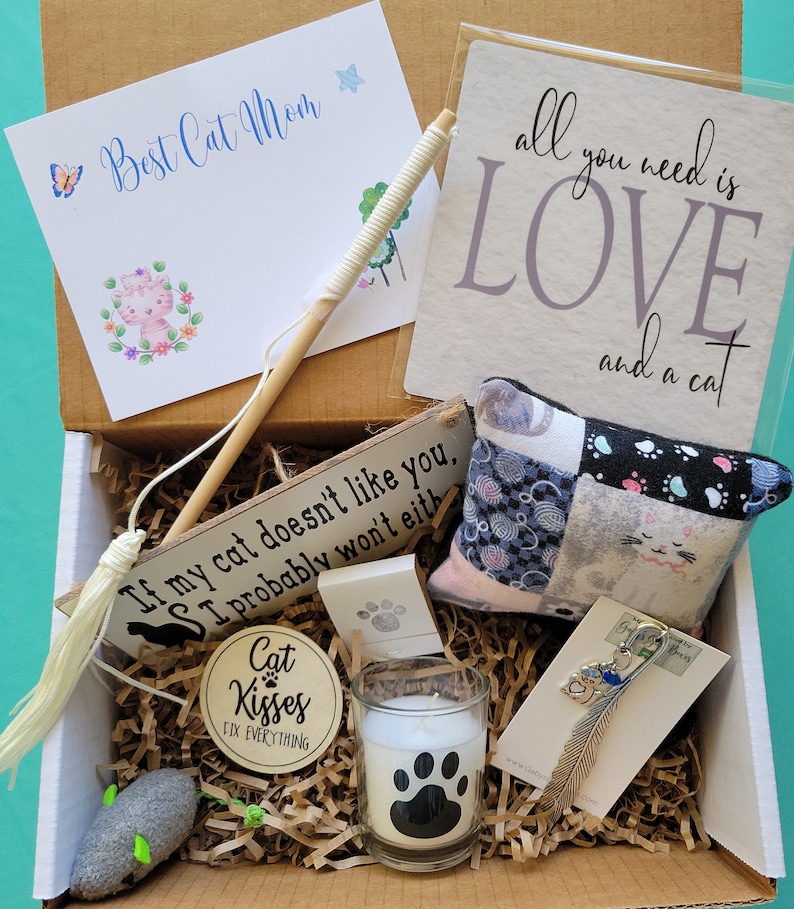May include: A brown cardboard box filled with cat-themed gifts. The box contains a wooden sign that reads 'If my cat doesn't like you, I probably won't either', a white candle with a black paw print, a small gray mouse toy, a white feather keychain with a cat charm, a small white and gray patchwork pillow with a cat design, a white wooden sign that reads 'Cat Kisses Fix Everything', and a white card with the words 'Best Cat Mom' in blue cursive writing.