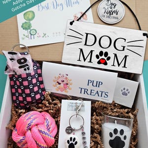 May include: A gift box containing a white wooden sign with the text DOG MOM and a paw print, a pink and black plaid waste bag dispenser, a neon pink rope ball, a keychain with a paw print and the name OAKLEY, a candle with a paw print, and treats.