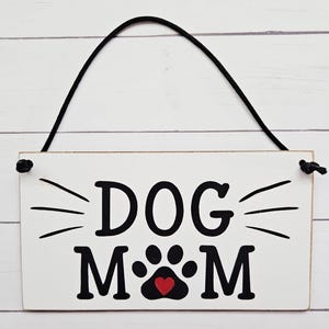 May include: White wooden sign with black text that reads "DOG MOM" with a black paw print with a red heart in the center. The sign has a black string for hanging.