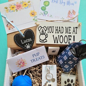 May include: A gift box filled with dog-themed items, including a blue ball, a dog bone keychain, a candle with a paw print, a bag of dog treats, a sign that says "You Had Me At Woof!" and a black chalkboard heart with the name "Lucas" written on it.