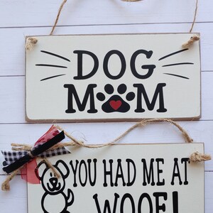 May include: Two wooden signs with black text and red hearts. The top sign says "DOG MOM" with a paw print in the middle. The bottom sign says "YOU HAD ME AT WOOF!" with a cartoon dog illustration.