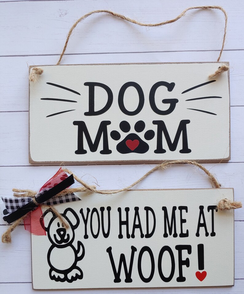 May include: Two wooden signs with black text and red hearts. The top sign says "DOG MOM" with a paw print in the middle. The bottom sign says "YOU HAD ME AT WOOF!" with a cartoon dog illustration.