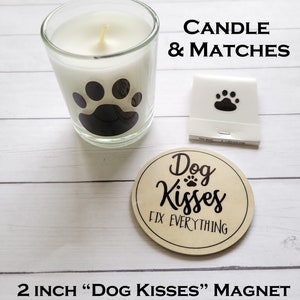 May include: A white candle with a black paw print design in a glass jar, a pack of matches with a paw print design, and a 2 inch round wooden magnet with the text "Dog Kisses Fix Everything" and a paw print.
