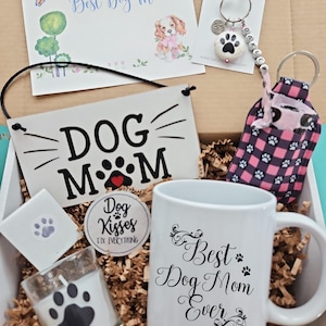 May include: A gift box filled with dog-themed items for a dog mom. The box includes a mug that says "Best Dog Mom Ever", a sign that says "Dog Mom", a candle with a paw print, a small bag with a paw print pattern, and a keychain with a paw print charm.