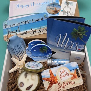 May include: A gift box containing a beach-themed collection: a sign reading The only BS I need is Beach and Sunshine, a Mrs can cooler, a keychain with charms, a candle with seashells, a luggage tag saying Safe Travels, and car coasters.