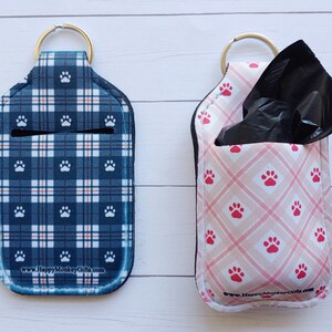 May include: Two keychains with paw print designs. The keychain on the left is blue and white plaid with white paw prints. The keychain on the right is pink with white paw prints and a black plastic bag dispenser. Both keychains have the text "www.HappyMonkeyGifts.com".