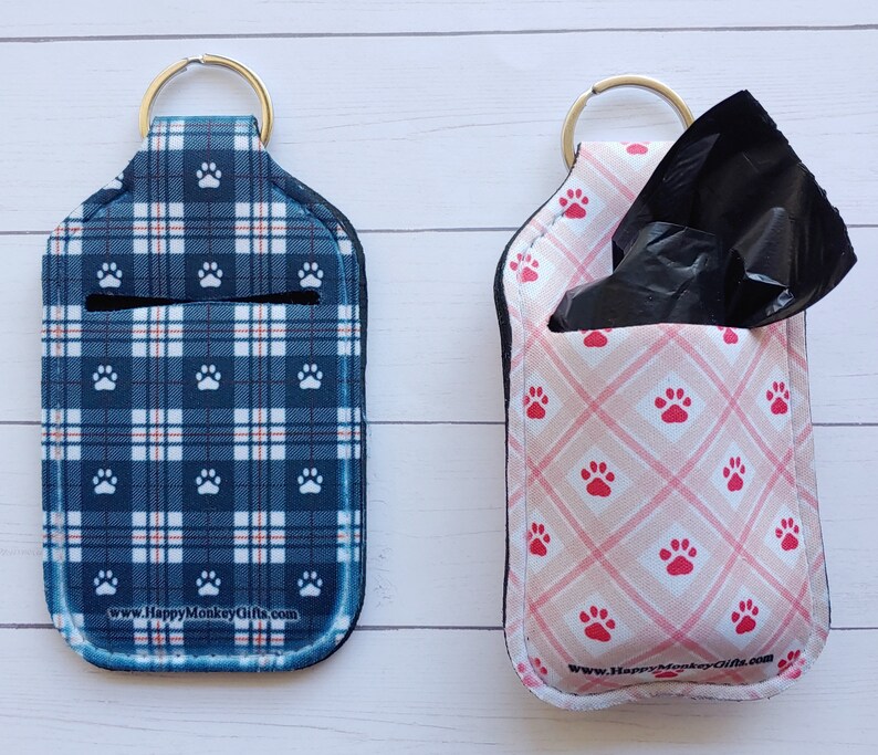 May include: Two keychains with paw print designs. The keychain on the left is blue and white plaid with white paw prints. The keychain on the right is pink with white paw prints and a black plastic bag dispenser. Both keychains have the text "www.HappyMonkeyGifts.com".