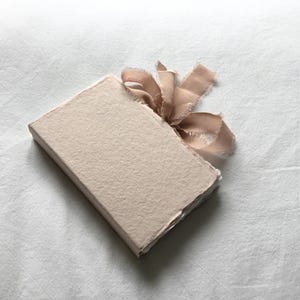 May include: A small, rectangular, light beige paper-covered box with a deckled edge, tied with a light brown ribbon. The box is resting on a white, textured surface. The ribbon is tied in a bow.