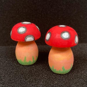May include: Two hand-painted ceramic mushroom figurines. The mushrooms have red caps with white polka dots and orange stems with green grass at the bottom.