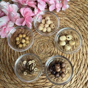 Op de afbeelding: Vijf doorzichtige glazen schaaltjes met diverse knutselkralen. Eén schaaltje bevat kleine lichte houten kralen, een ander grotere getextureerde houten kralen, één bevat gladde beige kralen, één donkerbruine kralen en de laatste kleine witte tussenkralen en metalen buisjes.