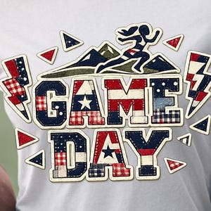 May include: White t-shirt featuring a "GAME DAY" design in red, white, and blue patchwork lettering. The graphic includes a runner silhouette, mountains, and lightning bolts. A brown football is held in the person's arms.