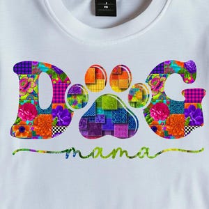 May include: White t-shirt featuring a colourful "Dog mama" design. The word "Dog" is in large, patchwork-style letters, with a paw print in the centre. The word "mama" is written in a cursive font below.
