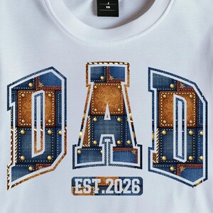 Könnte beinhalten: Weißes T-Shirt mit dem Wort "DAD" in einem Jeans- und Lederdesign, akzentuiert mit goldenen Nieten. Der Text "EST. 2026" befindet sich unter dem Wort "DAD". Das Shirt liegt gefaltet auf einer Holzoberfläche.