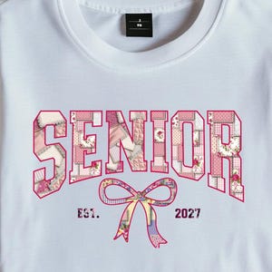 以下が含まれることがあります： ピンクのパッチワーク文字で「SENIOR」と書かれた白いTシャツ。 「SENIOR」の下には、ピンクと紫のリボンと「EST. 2027」の文字があります。 Tシャツは木製の表面に折りたたまれています。