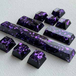 May include: A set of black and purple keyboard keycaps with a glittery finish. The keycaps are in various sizes and shapes, including a long space bar.