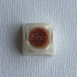 May include: A white keycap with a round, copper-colored insert. The insert has a textured, sparkly surface.