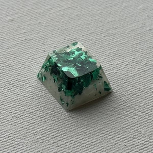 Puede incluir: Un keycap de resina verde y blanco con un acabado brillante. El keycap tiene forma de pirámide y una parte superior cuadrada.