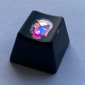 Puede incluir: Una tecla de teclado negra con un diseño de calavera iridiscente transparente.