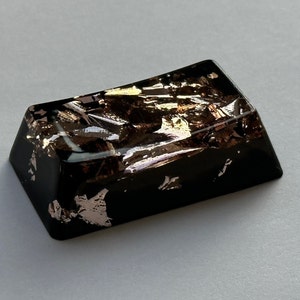 May include: A black keycap with a unique design featuring gold and pink flakes embedded in the resin. The keycap has a rectangular shape with rounded edges.