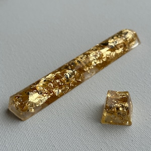 May include: Two gold resin keycaps with a gold leaf design. The larger keycap is rectangular and the smaller keycap is square.