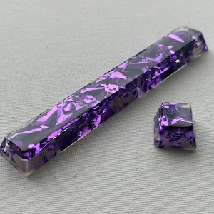May include: Two purple resin keycaps with a glittery finish. The larger keycap is rectangular and the smaller keycap is square.