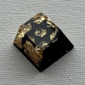 Op de afbeelding: Een vierkant zwart hars object met een bladgoud ontwerp. Het bladgoud is ingebed in de hars, waardoor een getextureerd, glinsterend effect ontstaat. Het object heeft een geometrische vorm en een glanzende afwerking.