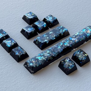 May include: A set of black resin keycaps with iridescent glitter inclusions. The keycaps are in various shapes and sizes, including a long space bar and several smaller keys. The glitter reflects blue, green, and purple hues.