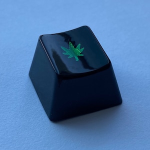 Puede incluir: Una tecla de teclado negra con un diseño de hoja de marihuana verde.