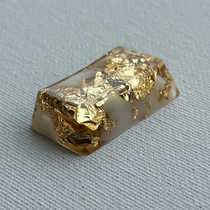 May include: A white resin keycap with gold leaf flakes embedded inside. The keycap is rectangular with rounded edges and a slightly raised surface.