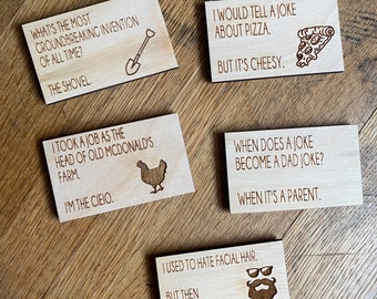 May include: Five wooden rectangular coasters with engraved jokes and illustrations. One coaster reads "What's the most groundbreaking invention of all time? The shovel." Other coasters have pizza, chicken, and beard graphics.