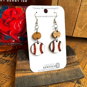 May include: A pair of baseball-themed earrings made from repurposed Kentucky bourbon barrels. The earrings feature a white baseball with red stitching and a brown wood circle.