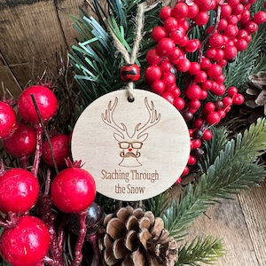 May include: Wooden Christmas ornament with a reindeer design and the text "Stashing Through the Snow". The ornament is brown with a red nose and black antlers. It is hanging from a red and black plaid ribbon.