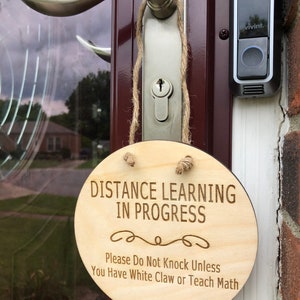 May include: A wooden sign with the text "Distance Learning in Progress" and the message "Please Do Not Knock Unless You Have White Claw or Teach Math".