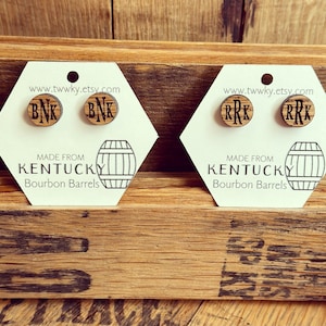 May include: Two pairs of wooden earrings with initials on a white card. The card says "Made from Kentucky Bourbon Barrels" and has a barrel illustration. The earrings are displayed on a wooden surface.