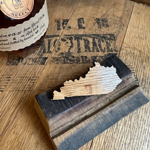 May include: A wooden state of Kentucky cutout on a wooden stand. The stand is made from a repurposed barrel stave. The state is a light brown color and the stand is a dark brown color.