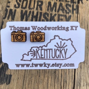 Puede incluir: Una tarjeta blanca con el texto "Thomas Woodworking KY" y un dibujo del estado de Kentucky con la palabra "KENTUCKY" en su interior. Dos pequeños pendientes de madera con forma de cámara están en la tarjeta. La dirección del sitio web "www.twwky.etsy.com" está impresa en la tarjeta.
