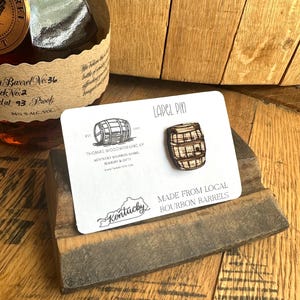 May include: A wooden lapel pin shaped like a bourbon barrel, displayed on a white card with text. The card reads "Made from Local Bourbon Barrels" and "Kentucky". A bottle of bourbon is in the background.