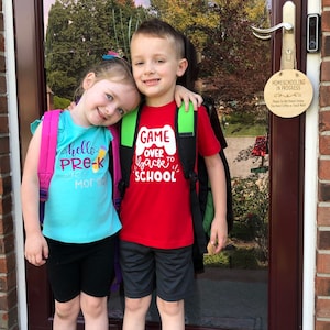 May include: Two children, a girl and a boy, stand in front of a door with a sign that reads "Homeschooling in Progress". The girl is wearing a turquoise shirt with the text "hello pre-k" and a pink backpack. The boy is wearing a red shirt with the text "Game Over Back to School" and a green backpack. They are both smiling.